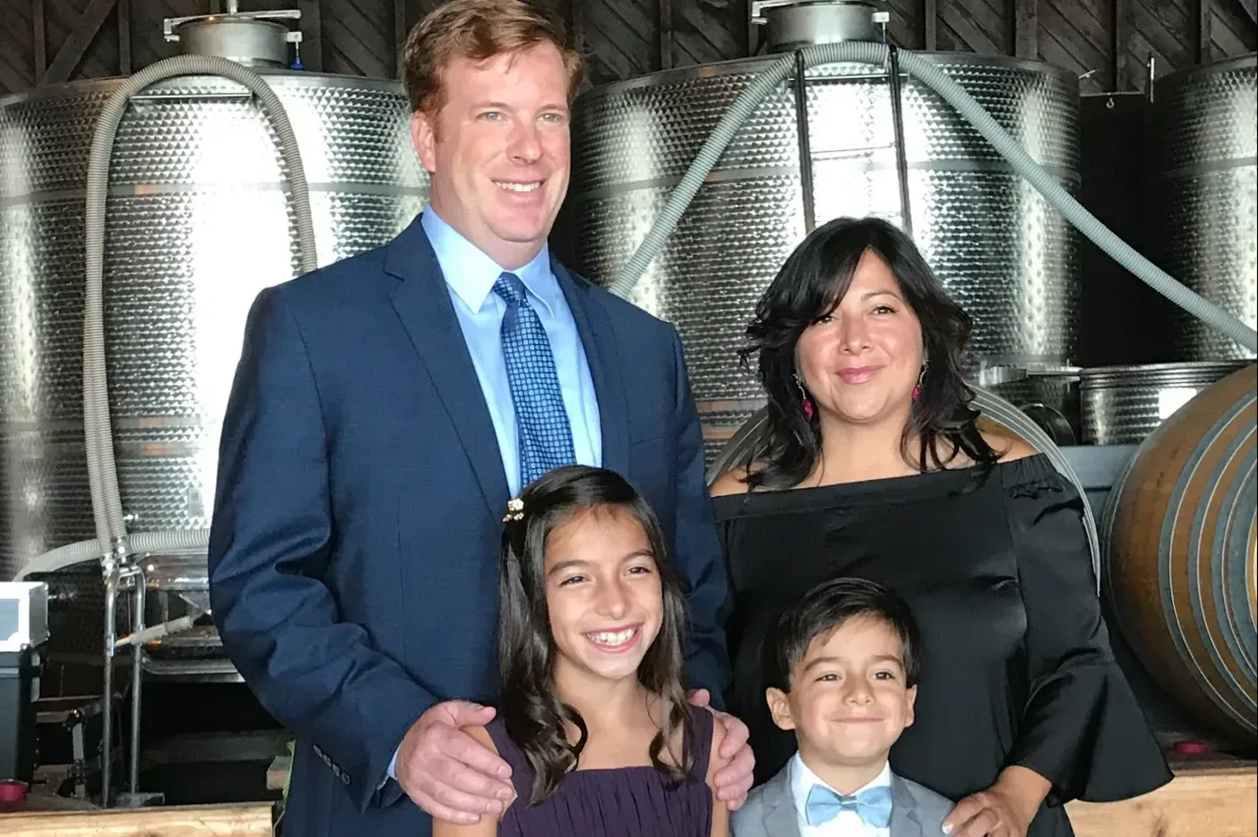 A man and woman stand behind a smiling girl in a purple dress and a boy in a gray suit and bow tie. They are in front of large metal wine casks inside a rustic building, likely a winery. The man is in a blue suit, and the woman is in a black off-the-shoulder dress.