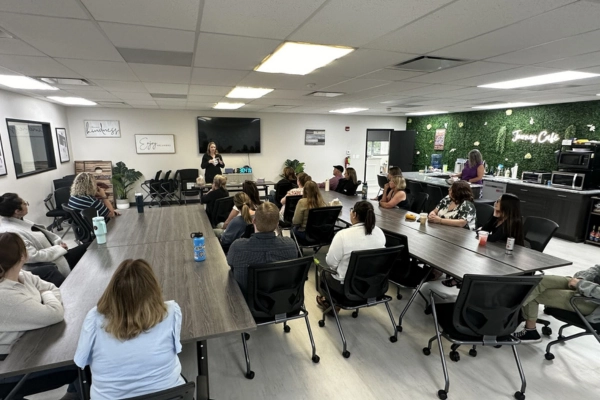 A group of people sit at tables in a modern, well-lit meeting room. They are facing a presenter standing at the front of the room with a TV screen behind her. The room features a well-stocked kitchenette with a green wall labeled "Jenny's Cafe.