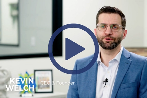 A man with glasses and a beard, wearing a blue suit and white shirt, is speaking in an office setting. "Kevin Welch, CEO & Founder," is written in the lower left corner. There is framed artwork in the background, and a play button icon is over the image.