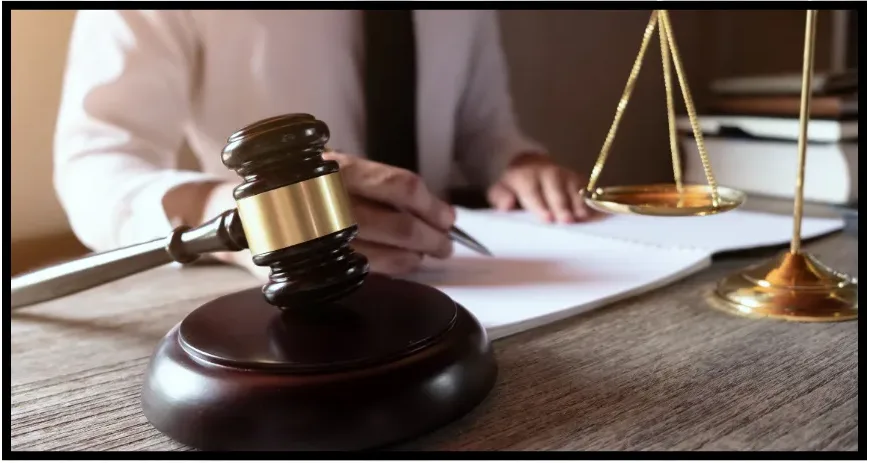 A judge or lawyer sits at a desk with a gavel and a justice scale in the foreground. The person is writing, wearing a white shirt and dark tie, with papers and books visible on the desk.