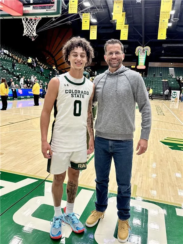 A basketball player in a Colorado State uniform stands on a court next to another person wearing a gray hoodie and jeans. The court is indoors, with banners and spectators in the background.